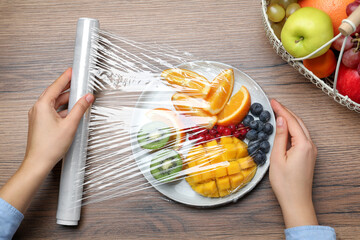 Wall Mural - Woman putting plastic food wrap over plate of fresh fruits and berries at wooden table, top view