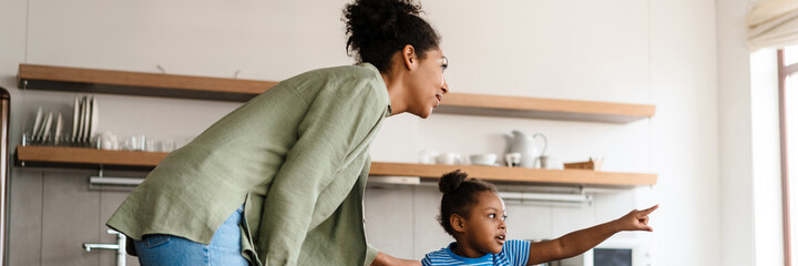 Wall Mural - Black happy mother and daughter pointing finger at at window while playing