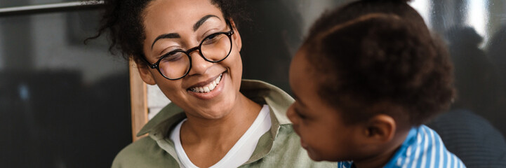 Black smiling woman playing her daughter while using mobile phone