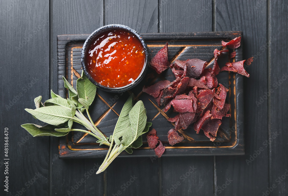 Board with spicy beef jerky and sauce on dark wooden background