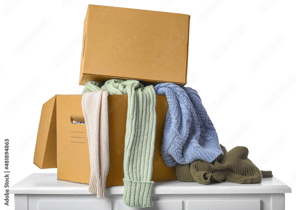 Cardboard boxes with different warm sweaters on chest of drawers against white background