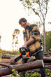 © Roman - bearded caucasian Worker in casual shirt and goggles cuts a felled tree trunk with powerful chainsaw. felled tree trunk is sawn by young lumberjack, at summer evening. Deforestation, forest cutting