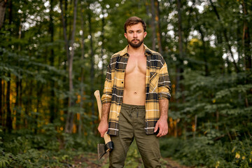 Wall Mural - Brutal handsome european man in open plaid shirt carry large splitting axe in summer forest natural landscape, lumberman with perfect muscular body posing at camera, shirtless sexy lumberman