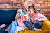 Caucasian happy family knitting. Senior woman and child girl, grandmother is teaching little girl to knit. Crafts for kids. Using Knitting tools, needles, have talk while sitting at home on sofa