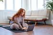 © Roman - Smiling fat woman waiting for online personal trainer, looking forward for home training. overweight caucasian woman sitting on fitness mat at home in living room, using laptop, in sportswear