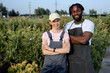 © Roman - portrait positive black man in garden or greenhouse, standing with redhead female. professional gardeners in apron working with blooming flowers in pots. Selective focus. Gardening activity and summer