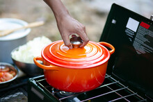 Man's Hand Putting Cover On A Red Enameled Cast Iron Dutch Oven On Camping Stove