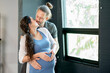 © narong - Pregnant woman and her husband hug and kiss together in front of glass window express love and care for family.