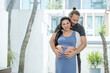 © narong - Man hug pregnant woman from back and woman look at camera and smiling. They stand in living room with concept of happiness family at home together.
