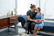 © narong - Caucasian husband and pregnant wife sit on sofa and hold red heart symbol and also kiss together to express love and care of their family in living room with glass windows.