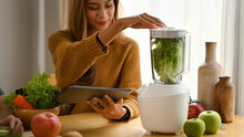Healthy Woman Using Digital Tablet And Making Green Vegetables Detox Smoothie In Kitchen.