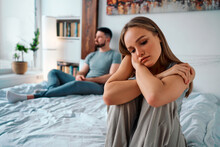 Upset Young Woman In Home Clothes Sitting On The Bed, In The Background Sitting Man. Family Troubles.