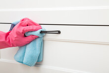 Woman Hand In Pink Rubber Protective Glove Using Blue Dry Rag And Wiping Black Metal Handle On White Wooden Dresser In Room. Closeup. Side View. Regular Cleanup At Home.