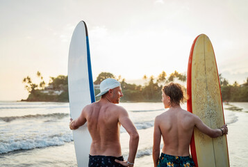 Wall Mural - Father with teenager son standing with surfboards on the sandy ocean beach with palm trees on background lightened with sunset sun. They smiling and have a conversation. Family active vacation concept