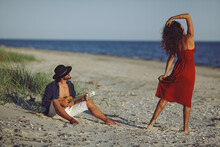 Happy Couple In Love Sensual Romance. Caucasian Man Playing Ukulele Serenading And Girl In Red Dress Dancing On The Beach At Sunset. A Bearded Handsome Man And Stunning Woman Having Romantic Tender