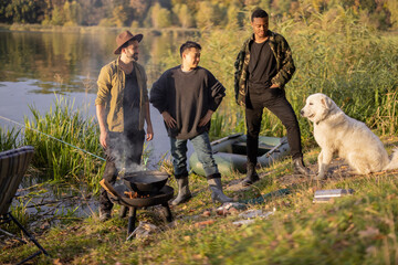 Multiracial group of male friends talking and cooking Ukha on fire. Men resting and fishing on lake coast at autumn. Leisure and weekend in nature. Friendship