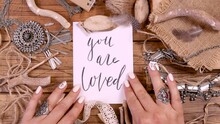 Top View Of A Young Woman Touching A Handwritten Note With Words YOU ARE LOVED On Wooden Table With Ethnic Jewelry And Natural Decor. Handmade Craft Accessories, Love, Valentine's Concept
