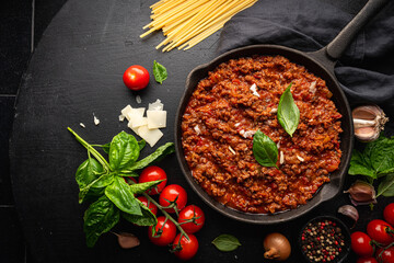 Wall Mural - classic italian bolognese sauce stewed in a pan with ingredients on black tile background, top view