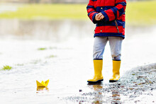 Happy Little Kid Boy In Yellow Rain Boots Playing With Paper Ship Boat By Huge Puddle On Spring Or Autumn Day