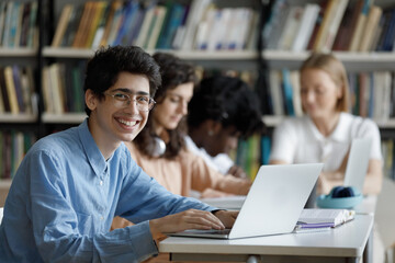 Happy gen Z high school student in glasses working on study project at laptop in library, watching online learning presentation, virtual class, lesson, sitting by classmates. Head shot portrait