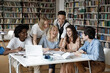 © fizkes - Happy joyful mixed race group of students working in library together, talking at table with books and laptop, laughing, having fun, discussing research study project. Education concept