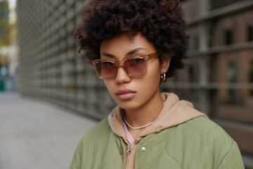 Wall Mural - Serious stylish woman with curly hair wears sunglasses jacket and hoodie looks directly at camera walks outdoors during daytime stands against blurred background. People and lifestyle concept