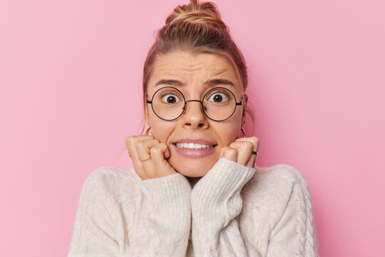 Photo of anxious stressed woman clenches teeth looks with frightened expression keeps hands uder chin reacts on something horrible wears round spectacles and sweater poses against pink wall.
