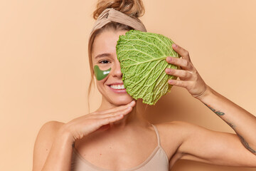 Wall Mural - Tender young woman touches chin gently smiles broadly covers face with cabbage leaf applies green hydrogel patches wears headband and t shirt isolated over beige background. Sin care concept