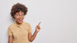 © WHstudio Leushin N - Attractive smiling young curly woman points index fnger at upper right corner demonstrates promotio gives way somewhere wears round transparent glasses and t shirt isolated over white background.
