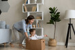 © fizkes - Joyful loving millennial caring mother on nanny pushing cardboard box with seated small laughing child daughter, running barefoot in heated warm wooden floor, playing entertaining game at home.