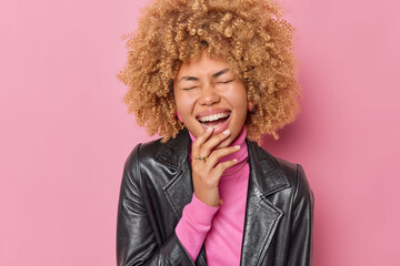 Wall Mural - Portrait of overjoyed curly haired woman laughs out joyfully keeps eyes closed hears something very funny wears leather jacket isolated over pink background. People emotions and style concept