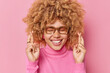 Hopeful cheerful young woman keeps fingers crossed believes in good luck makes wish anticipates something smiles broadly wears spectacles and casual turtleneck isolated over pink background.