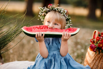 Wall Mural - picnic with family. cute little girl eating big piece of watermelon on straw stack in summertime in the park. Adorable child wearing in flowers wreath on head and blue dress