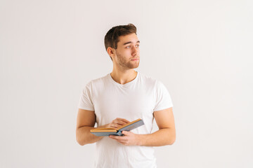 Wall Mural - Studio portrait of focused young man holding in hands paper book on white isolated background in studio, looking away. Front view of serious male student studying reading educational materials.