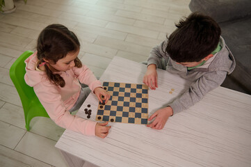 Overhead view of two adorable Caucasian elementary aged kids, boy and girl, brother and sister having great time playing checkers board game together at home interior.