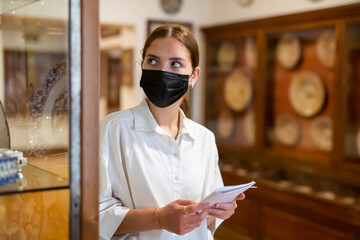 Young attractive girl in face mask holding brochure in archaeological museum and looking at ceramic exhibits