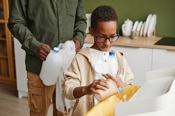 Canvas Print - Portrait of young African-American boy sorting waste at home with father in background, copy space