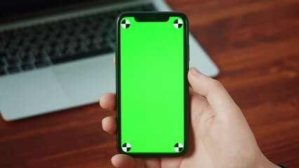 Businessman using smartphone with chroma key close-up. Young student holding mobile phone with green screen display, sitting at table with laptop computer.