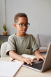 © Seventyfour - Vertical portrait of smiling African-American boy wearing glasses using laptop while studying at home, homeschooling concept
