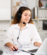 Young positive woman customer support phone operator in microphone headset working on laptop in the office