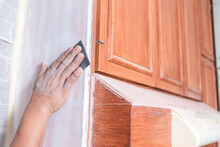 A Handyman Using A Piece Of Sandpaper To Smoothen Out The Edge Of A Wall Cabinet Prior To Painting. Home Renovation Or Finishing Works.