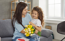 Young Caucasian Woman Enjoys Love, Presents And Flowers From Her Child On Mother's Day. Happy Mom Sitting On Couch At Home Hugs Cute Little Daughter And Thanks Her For Postcard And Bouquet Of Tulips