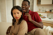 © Anatoliy Karlyuk - Selective focus of serious adult attractive charming woman with discontent face expression sitting on couch with african guy trying to get her attention, making faces behind her back, joking