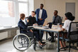 © Seventyfour - Wide angle view at diverse group of business people at meeting table in office with woman using wheelchair in foreground, copy space
