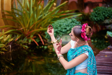 Beautiful young caucasian woman in traditional indian clothing sari with bridal makeup and jewelry and henna tattoo on hands dancing in temple garden.