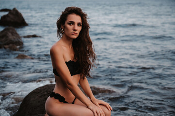 Wall Mural - young girl in a black swimsuit is sitting sexy on the rocks on the beach by the sea in summer on vacation