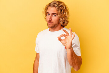 Wall Mural - Young caucasian man isolated on yellow background winks an eye and holds an okay gesture with hand.