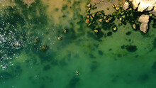 Aerial View Of A Girl Swimming In Sea With Algae Green Water By The Rocky Beach. Coast Of Sea In Summer. Top View. Landscape With Clear Azure Water, Stones And Rocks. Nature Background