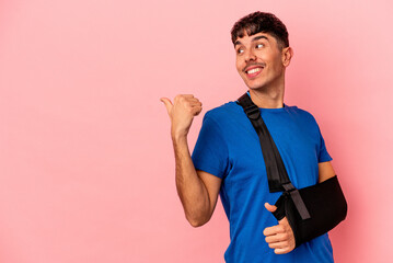 Wall Mural - Young mixed race man with broken hand isolated on pink background points with thumb finger away, laughing and carefree.