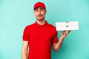 Young delivery man taking pizzas isolated on blue bakcground dreaming of achieving goals and purposes